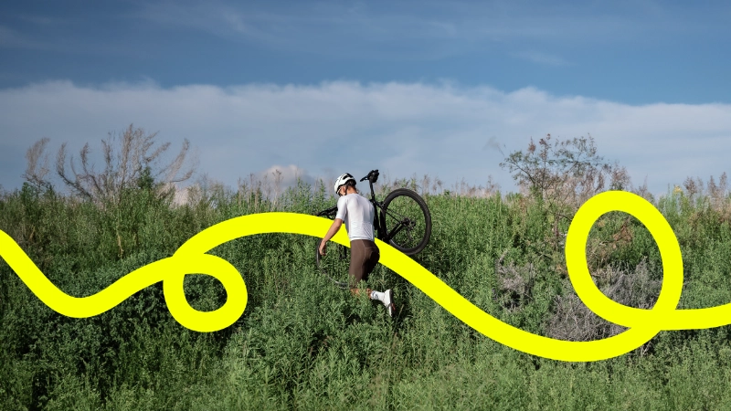 Radler trägt sein Gravelbike durchs Gestrüpp, eine gelbe JobRad Loop Linie kringelt sich durchs Bild