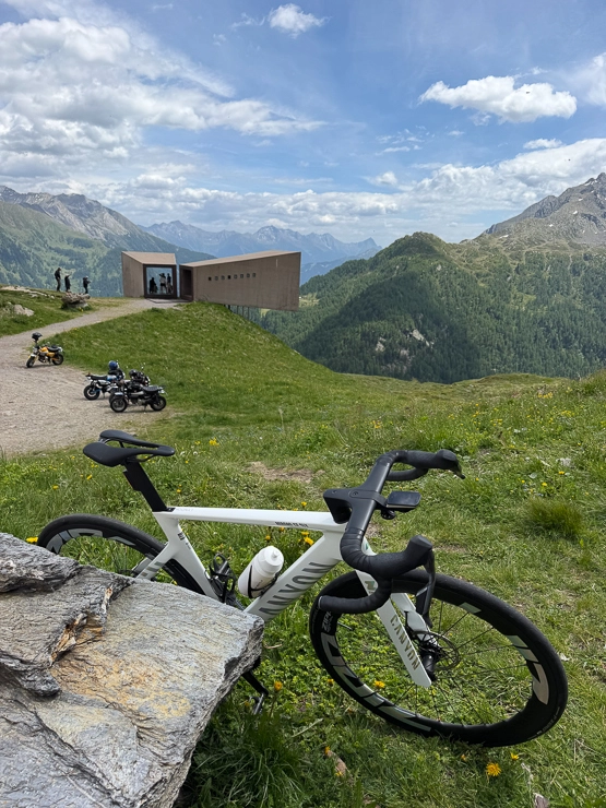 Weißes Rennrad auf einer grünen Bergwiese mit Alpenpanorama, modernes Gebäude und Motorräder im Hintergrund unter blauem Himmel.