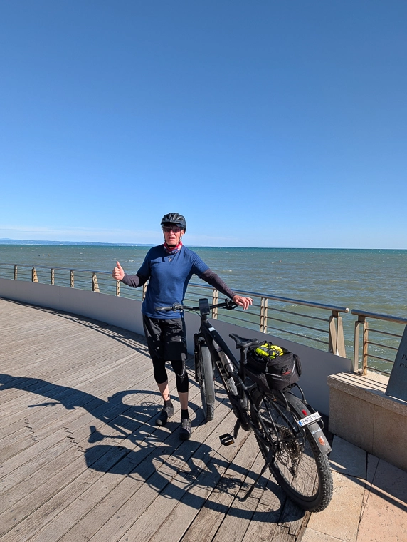 Eine Person steht mit einem E-Bike auf einer Uferpromenade am Wasser und zeigt mit erhobenem Daumen in die Kamera.
