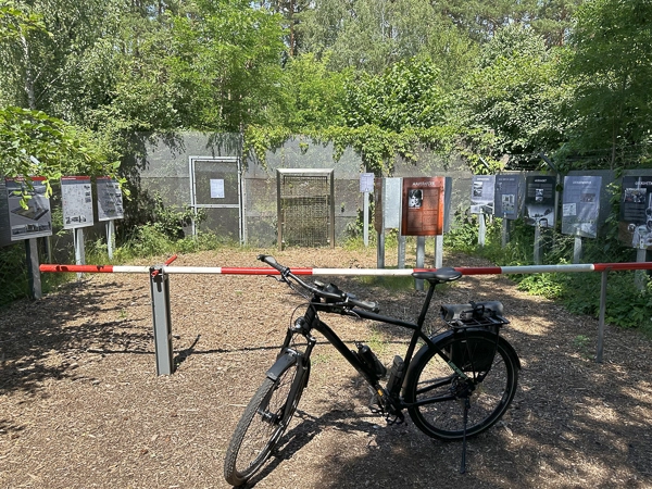 Fahrrad vor einer abgesperrten historischen Gedenkstätte im Wald, mit Informationstafeln und überwucherten Betonmauern im Hintergrund.