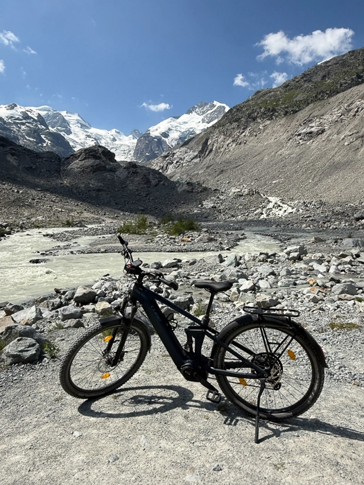Schwarzes E-Bike auf steinigem Gelände vor einem Gletscherfluss und schneebedeckten Bergen unter blauem Himmel.